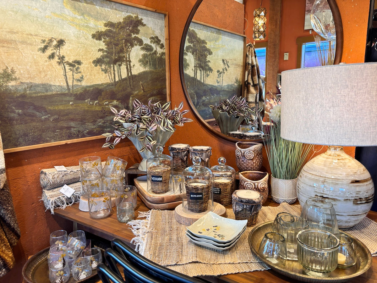 Autumn tabletop display at Over the Fence with a landscape painting, round mirror, candles under glass cloches, owl planters, flower-etched glassware and a ceramic table lamp