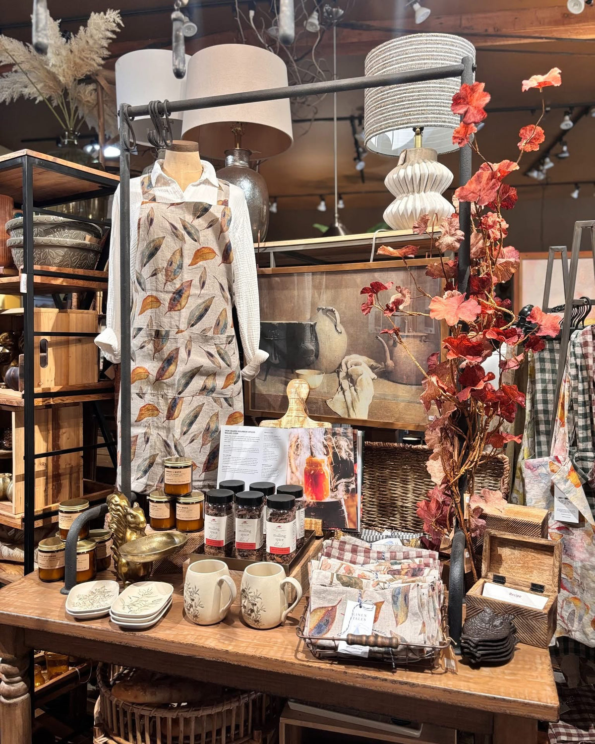 Fall kitchen display with a leaf-print linen apron, mulling spice, candles, botanical mugs and dishes, and table lamps