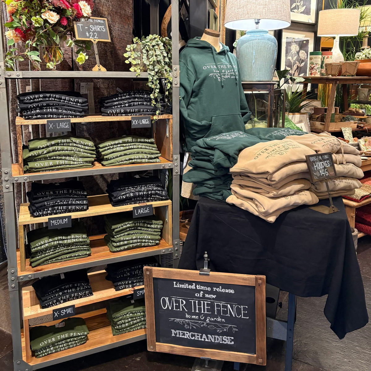 Over the Fence T-shirts and hoodies in black, green and tan on shelves, with a chalkboard sign announcing new Over the Fence merchandise